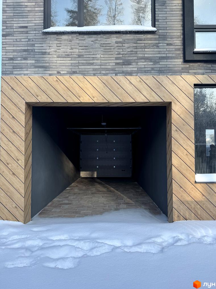 Вхід до гаража житлового комплексу Таунхауси Madera House з дерев'яними елементами та цегляною стіною. Прибудинкова територія засніжена.