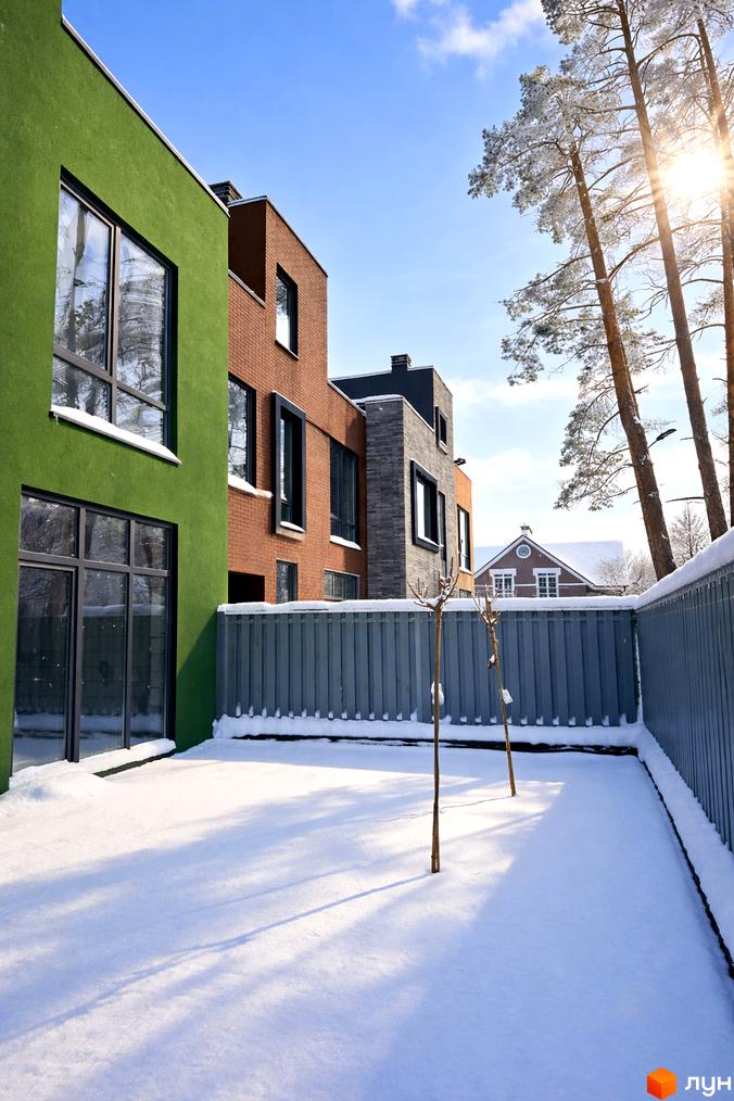 Таунхауси Madera House з зеленим та цегляним фасадом на тлі зимового двору з деревами та парканом. Сонячний день.