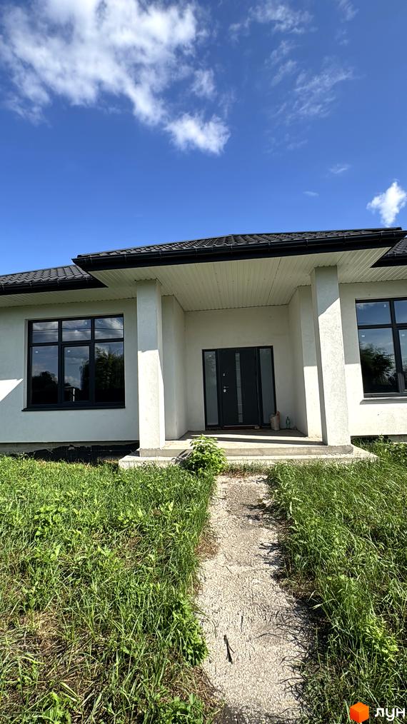 Вхідна група котеджу з великими вікнами, навколо зелена територія з травою та стежкою.