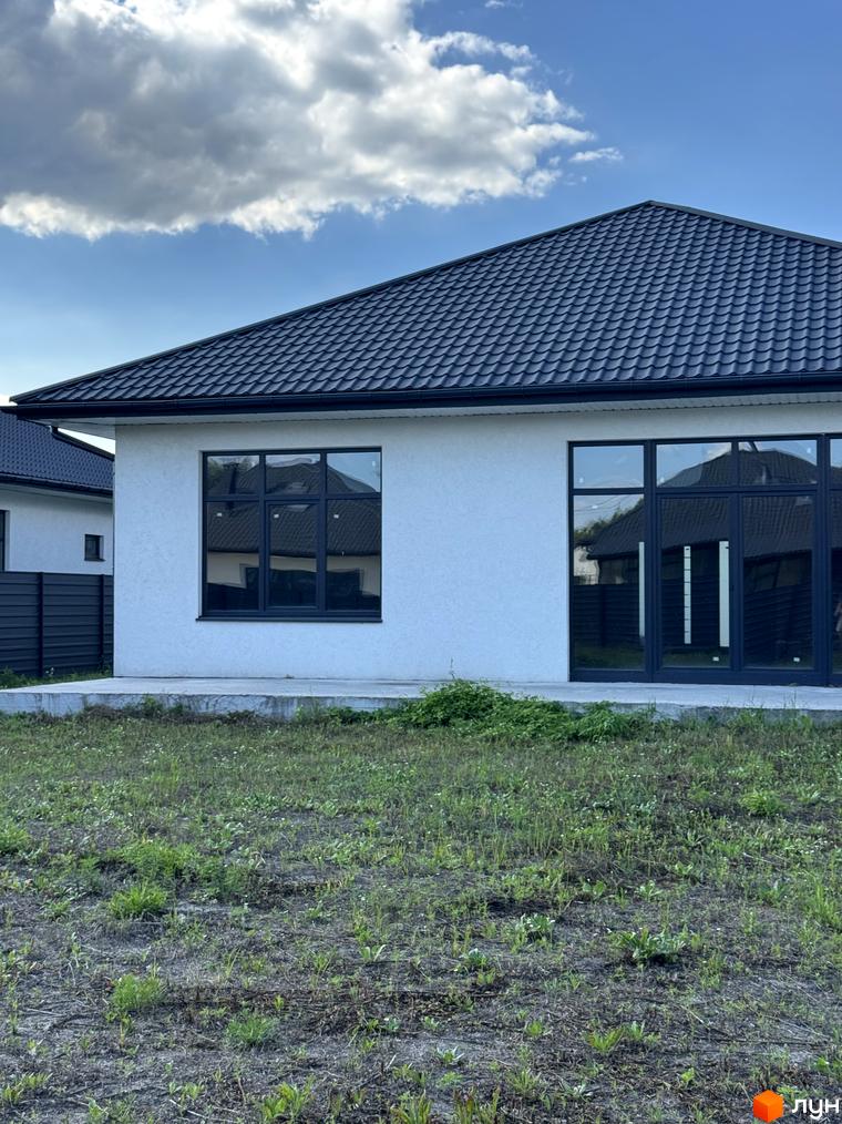 Зовнішній вигляд котеджу з великими вікнами та дахом, прибудинкова територія з невисокою рослинністю.