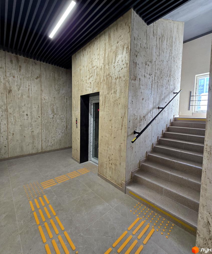 Внутрішній вигляд житлового комплексу ЖК КОРОНА з ліфтом та сходами, оформленими в світлих тонах. Простора зона з плиткою та тактильними елементами на підлозі.