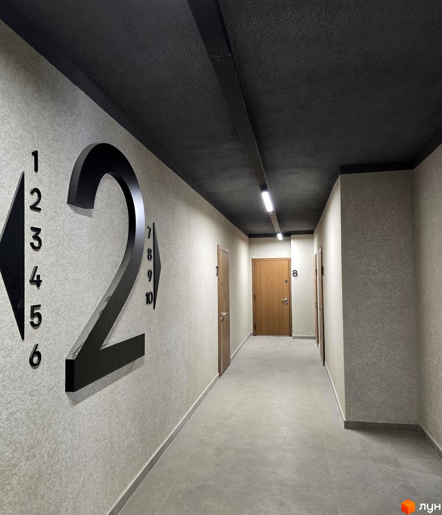 Коридор житлового комплексу ЖК КОРОНА з номерними знаками та дверима квартир. Світлі стіни та підлога з плитки створюють просторий вигляд.