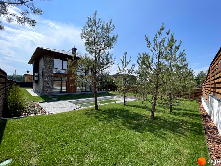 Зовнішній вигляд котеджного комплексу КМ Нова Ходосівка з великими вікнами та прибудинковою територією з газоном і деревами.