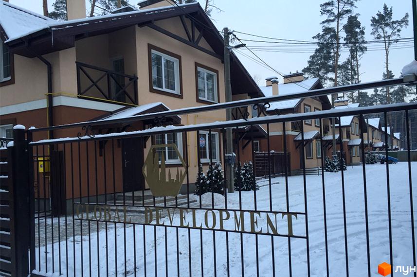 Будинки котеджного містечка Sweet Home мають бежево-коричневі фасади та темні дахи, вкриті снігом. Територія вкрита снігом, видно металеву огорожу та сосни.