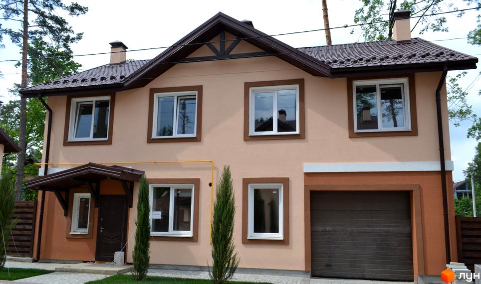 Фасад двоповерхового будинку Котеджного містечка Sweet Home з бежевими стінами, коричневим дахом і гаражем. Перед будинком вимощена доріжка та молоді хвойні дерева.