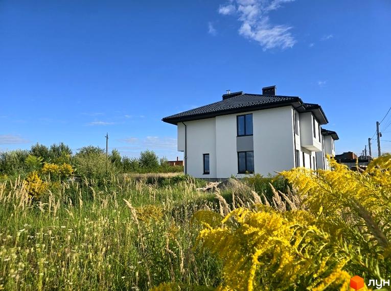 Білий двоповерховий будинок з темним дахом та вікнами в Котеджному містечку Savanna. Навколо висока трава та жовті квіти під блакитним небом.