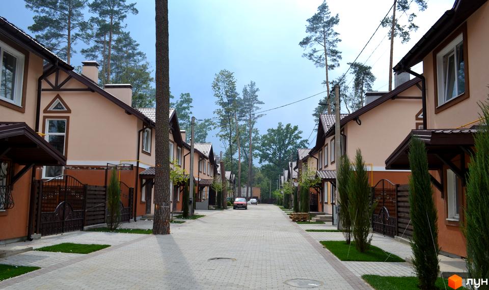 Будинки котеджного містечка Sweet Home мають світлі фасади та коричневі дахи. Вулиця вимощена бруківкою, з зеленими газонами та високими деревами.