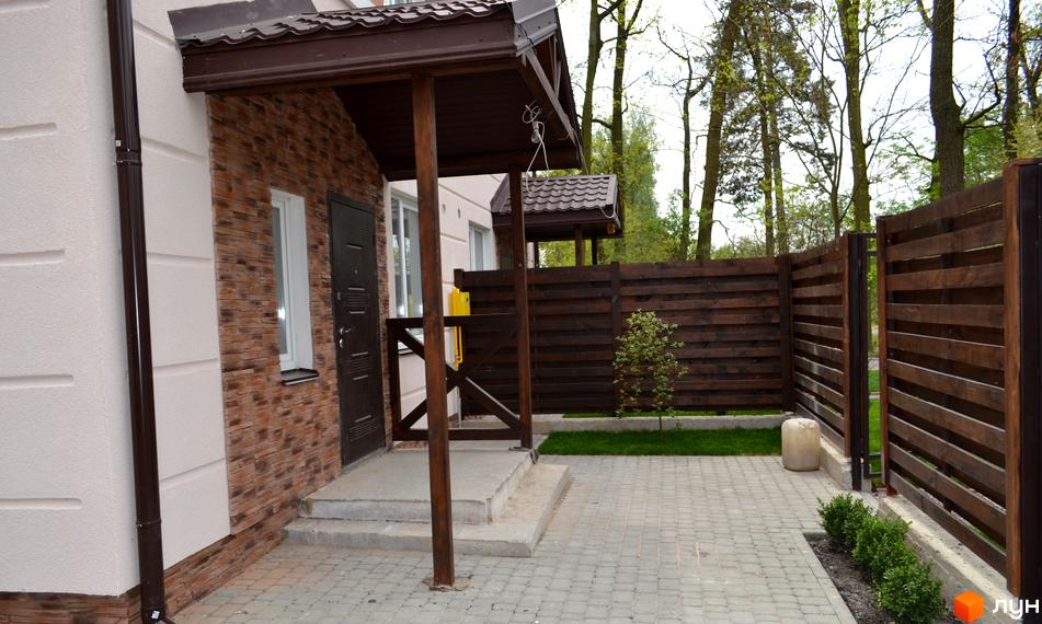 Будинок Котеджного містечка Sweet Home зі світлими стінами та коричневою обробкою. Прибудинкова територія має вимощення, газон та дерев'яний паркан.