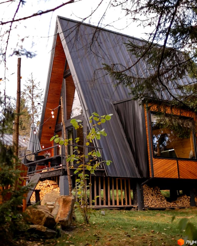 Котеджне містечко Yahody. Дерев'яний будинок A-frame з темним дахом, балконом та дровами під ним, розташований серед зелені та дерев.