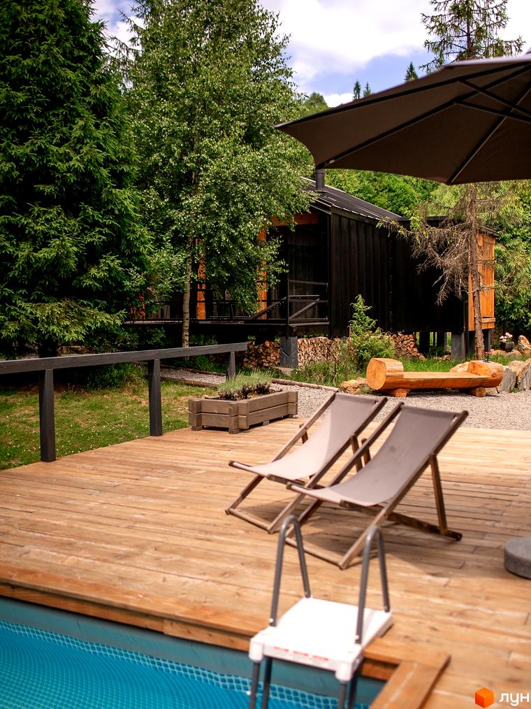 Чорний дерев'яний будинок на палях оточений деревами. Навколо будинку тераса з басейном, шезлонгами та парасолькою в Котеджному містечку Yahody.