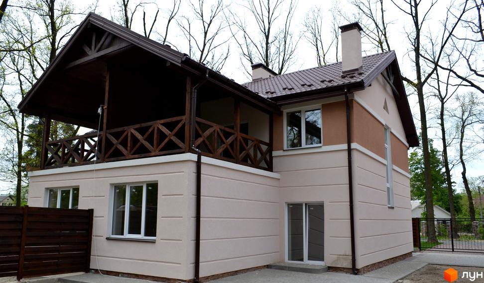 Котеджне містечко Sweet Home: двоповерховий будинок з бежево-коричневим фасадом, темним дахом та дерев'яним балконом. Навколо видно дерева та огорожу.