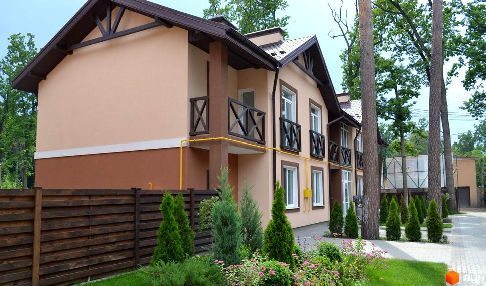 Котеджне містечко Sweet Home: двоповерховий будинок з бежевим фасадом, коричневим дахом та дерев'яними балконами. Прибудинкова територія з зеленими насадженнями.