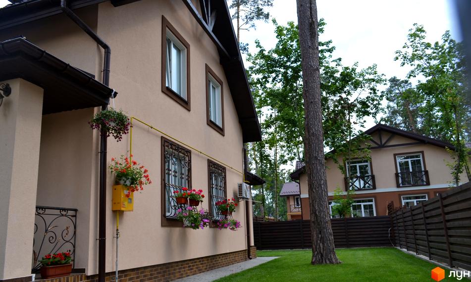 Фасад будинку Котеджного містечка Sweet Home з бежевими стінами, коричневими вікнами та дахом. Навколо доглянутий зелений газон та дерева.