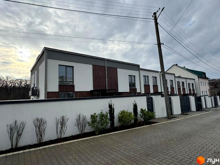 Таунхаус Luxor Gardens з білою стіною та дерев'яними вставками, перед ним доглянута прибудинкова територія з кущами та бруківкою.