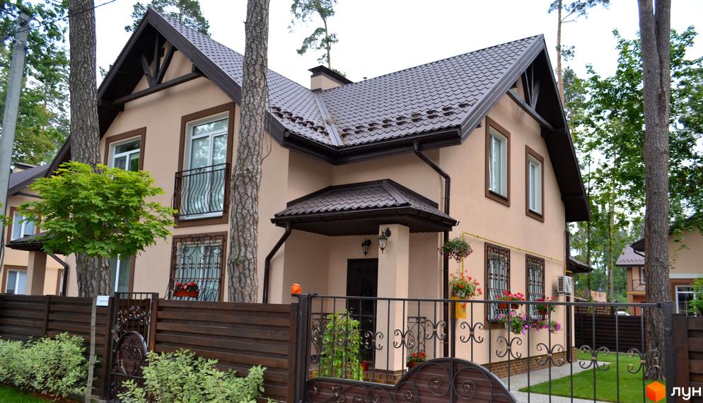 Будинок котеджного містечка Sweet Home з бежевими стінами, коричневим дахом, балконом та декоративними ґратами на вікнах. Прибудинкова територія має огорожу та дерева.