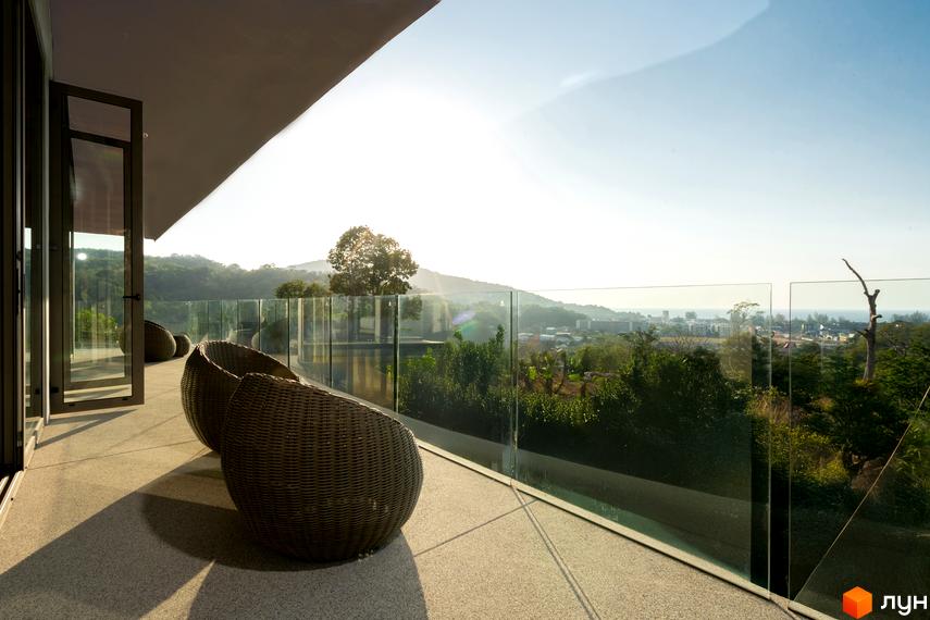 Балкон з плетеними кріслами та скляним огородженням у ЖК Botanic Club House, що відкриває вид на зелені пагорби та море.