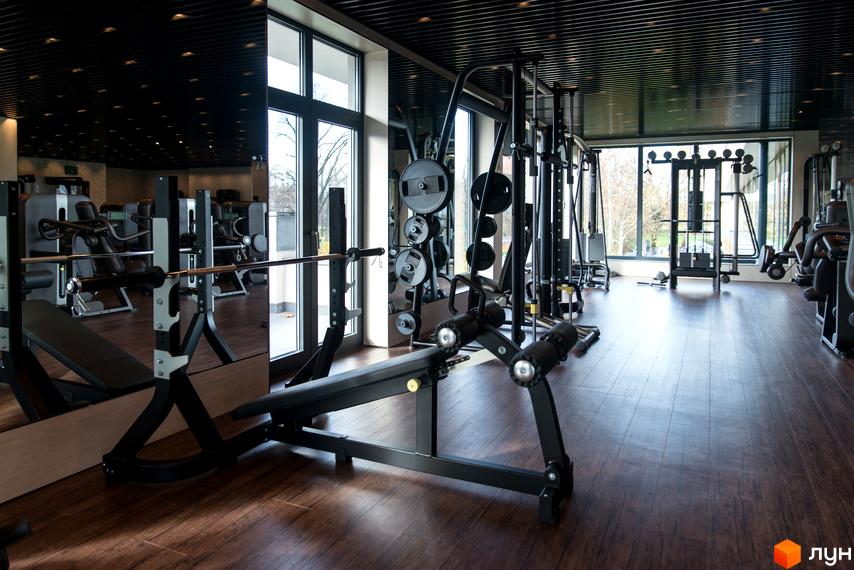 Сучасний тренажерний зал з різноманітним обладнанням для силових тренувань. Великі вікна забезпечують природне освітлення та вид на зелену територію.