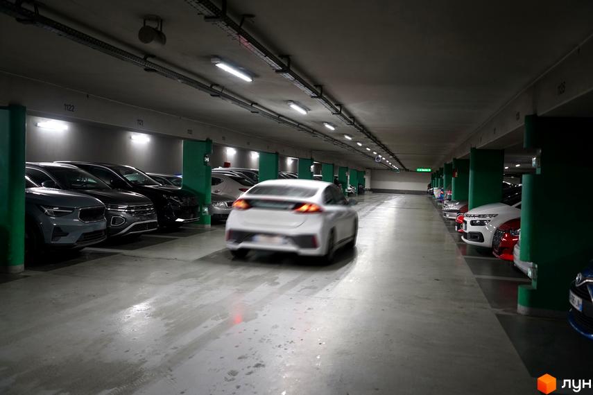 Підземний паркінг у житловому комплексі Botanic Club House з автомобілями, що паркуються. Паркінг має бетонну стелю та колони, освітлений люмінесцентними лампами.