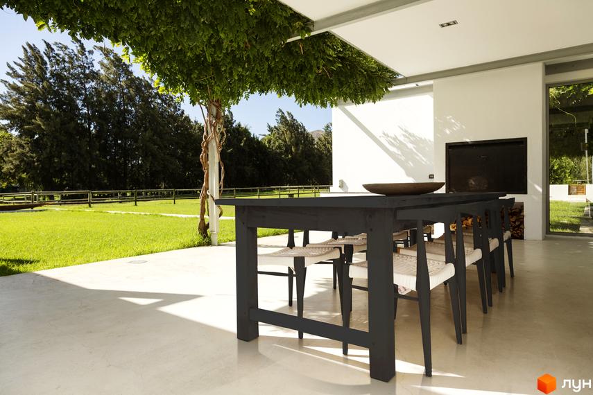 Відкрита тераса Житлового комплексу Botanic Club House з білими стінами, темним столом та видом на зелений газон і дерева.