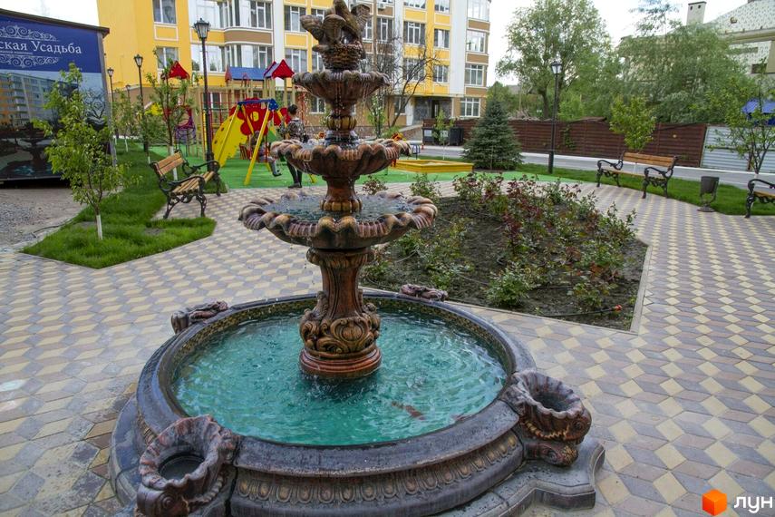Житловий комплекс Єнісейська садиба: жовтий багатоповерховий будинок з фонтаном та дитячим майданчиком на прибудинковій території.