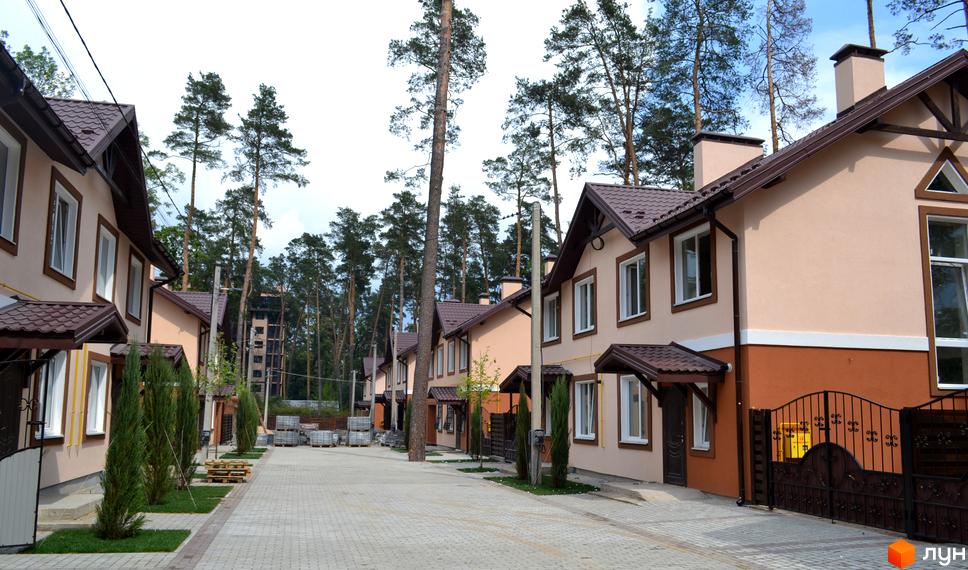 Котеджне містечко Sweet Home: двоповерхові будинки з бежевими фасадами та коричневими дахами. Прибудинкова територія вимощена бруківкою, є зелені насадження та сосни.