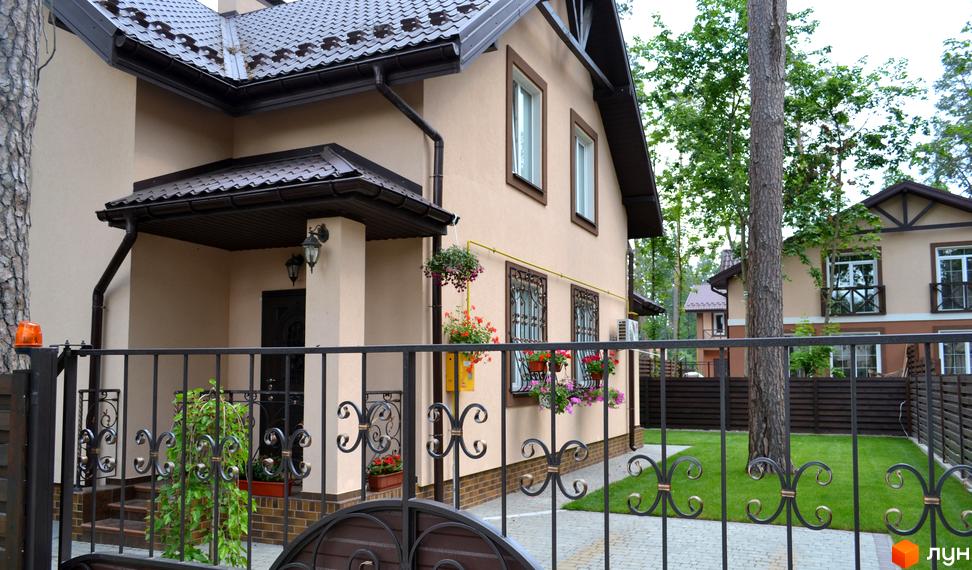 Фасад будинку Котеджного містечка Sweet Home з бежевими стінами, коричневим дахом та вікнами з декоративними решітками. Прибудинкова територія має зелений газон та кований паркан.