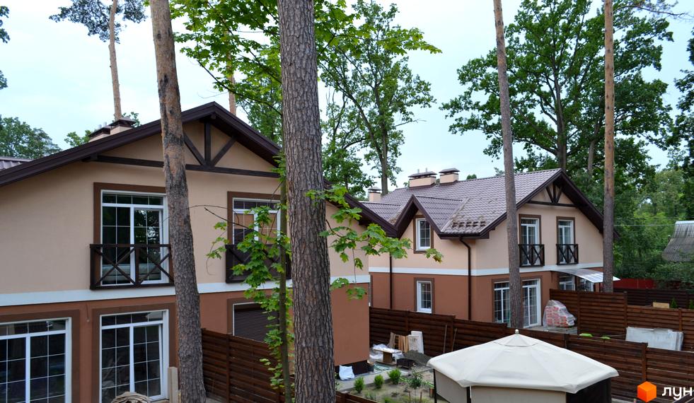 Двоповерхові будинки Котеджного містечка Sweet Home з бежевими та коричневими фасадами та коричневими дахами. Територія оточена деревами та дерев'яним парканом.