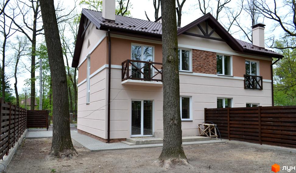 Двоповерховий будинок Котеджного містечка Sweet Home з бежево-коричневим фасадом та коричневим дахом. Навколо будинку є дерева та дерев'яний паркан.