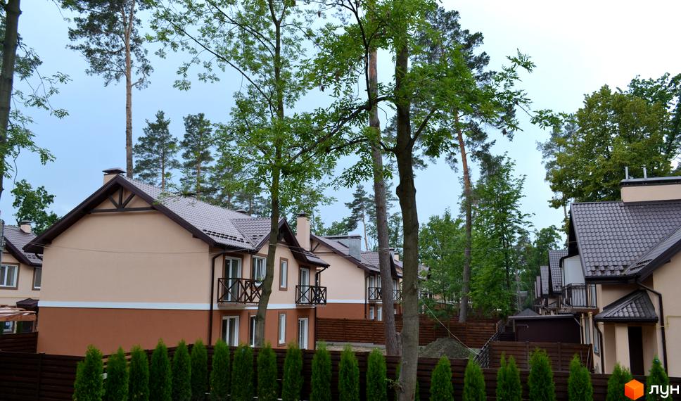 Двоповерхові будинки Котеджного містечка Sweet Home з бежево-коричневими фасадами та коричневими дахами. Навколо високі дерева та зелені кущі вздовж огорожі.