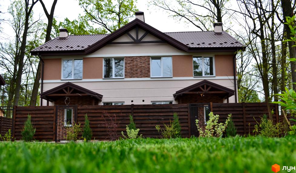 Двоповерховий будинок з коричневим дахом та світлими стінами в Котеджному містечку Sweet Home. Перед будинком зелений газон та дерев'яний паркан.