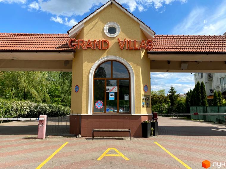 Житловий комплекс Гранд Віллас: жовта будівля з червоним черепичним дахом та арковим вікном. Перед в'їздом - паркан, лавка та паркувальні лінії.