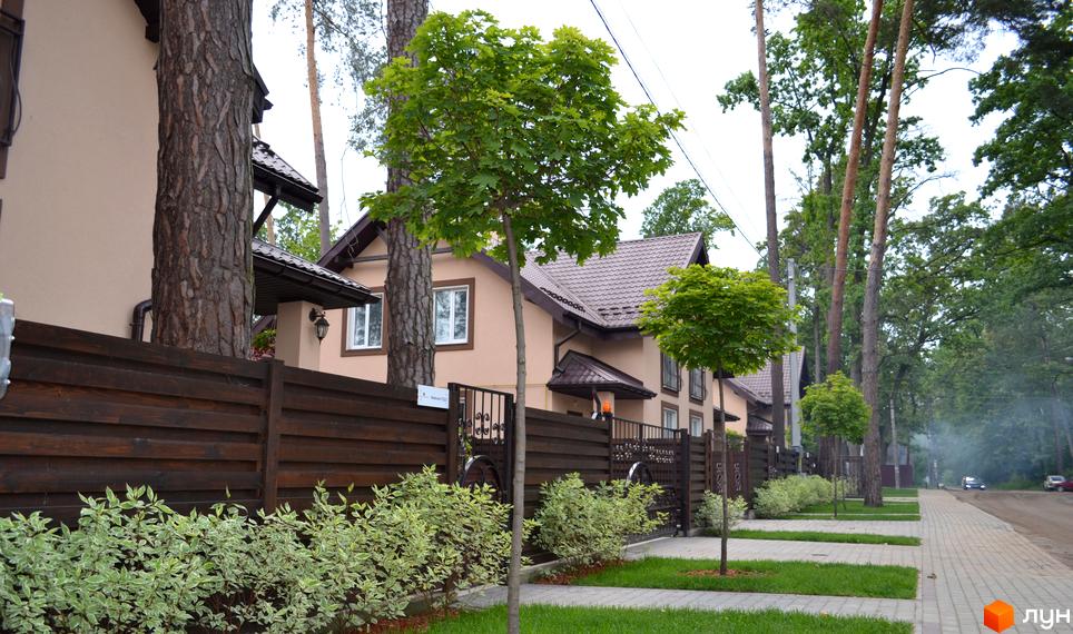 Фасади котеджів Котеджного містечка Sweet Home світло-бежевого кольору з коричневими дахами. Перед будинками розташовані дерев'яний паркан, зелені насадження та брукована доріжка.