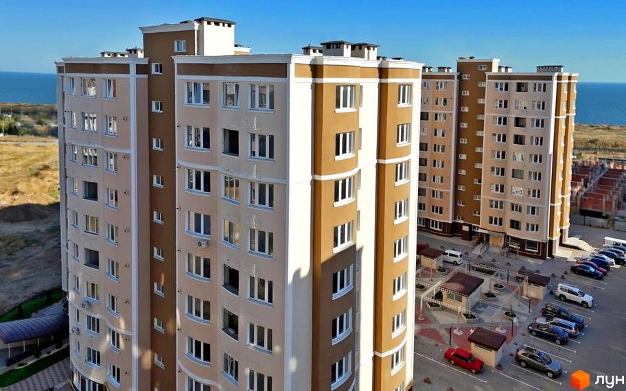 Житловий комплекс Дім у моря з коричневими та бежевими фасадами, вікнами та балконами, поруч розташовані парковка та дитячий майданчик.