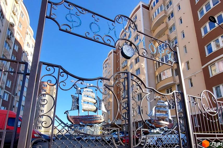 Житловий комплекс Дім у моря: ковані ворота з кораблями та годинником на тлі багатоповерхових будинків.