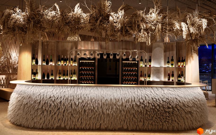 Інтер'єр бару в Апарт-готель IKON Resort з барною стійкою, оздобленою пухнастим матеріалом, та полицями з вином. Декорації з сухих рослин на стелі створюють затишну атмосферу для відпочинку мешканців новобудови.