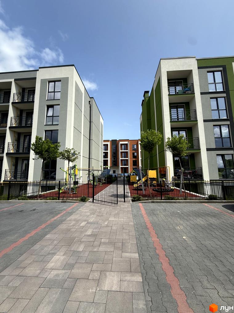 Житловий комплекс Brickwood 2: багатоповерхові будинки світло-сірого та зеленого кольорів з балконами. Перед будинками розташований дитячий майданчик та вимощена територія.