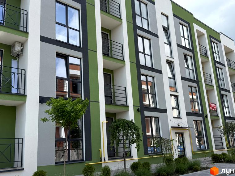 Житловий комплекс Brickwood 2 з фасадом у зелених, сірих, білих тонах, з вікнами та балконами. Перед будинком висаджені дерева та кущі.