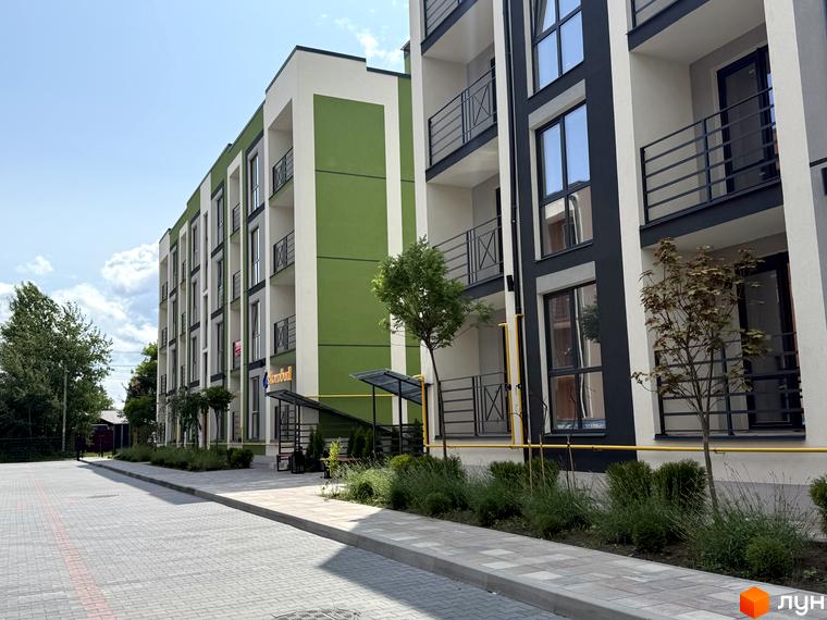 Фасад Житлового комплексу Brickwood 2 з білими, зеленими та темно-сірими секціями, балконами та вікнами. Перед будинками вимощена доріжка, невеликі дерева та кущі.