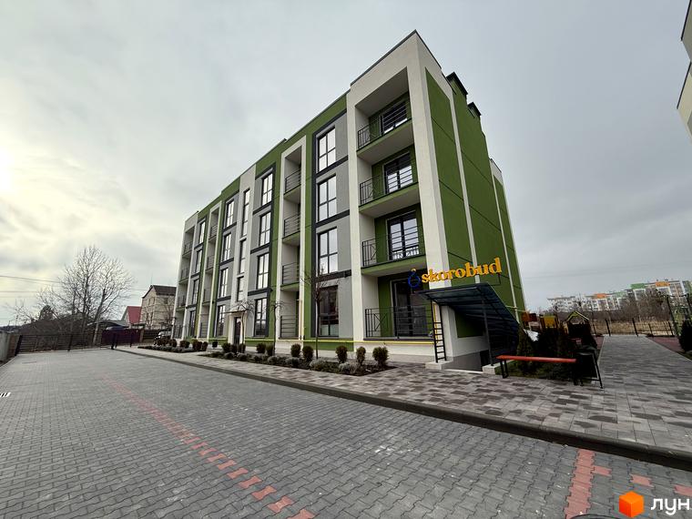 Зовнішній вигляд будинку ЖК Brickwood 2 з зеленими та білими фасадами, балконами та вхідною групою. Прибудинкова територія з бруківкою та зеленими насадженнями.