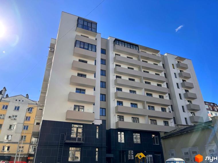 Житловий комплекс на вул. Володимира Великого, 16: багатоповерховий будинок зі світлим фасадом, темним цоколем та балконами. На території є дитячий майданчик.