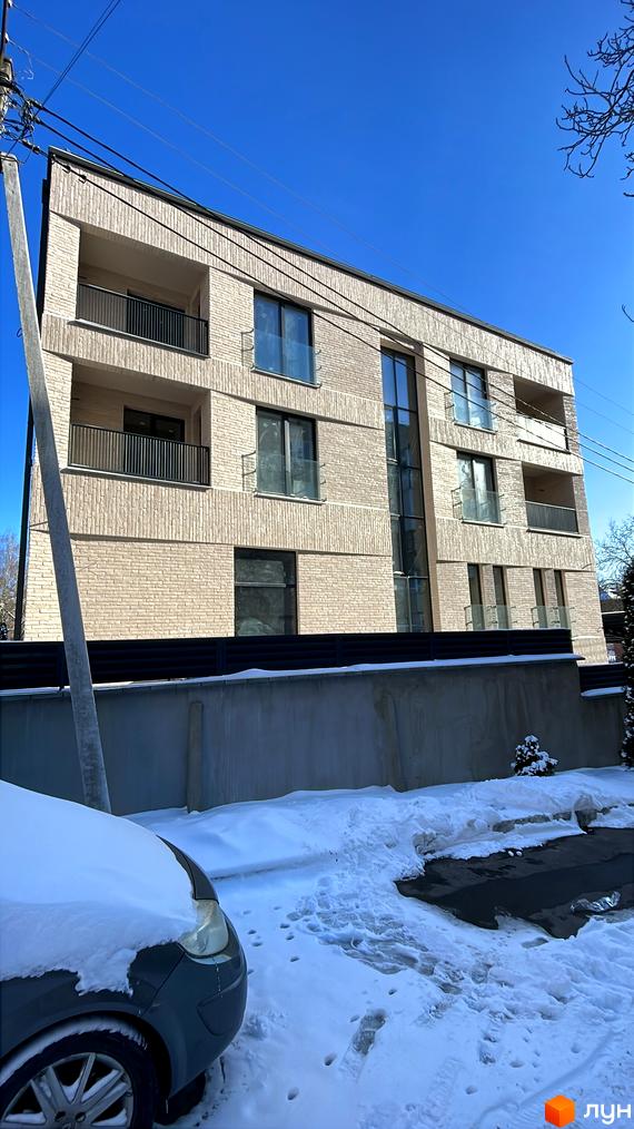 Багатоквартирний будинок з фасадом зі світлої цегли, балконами та великими вікнами. Прибудинкова територія вкрита снігом, видно парковку та огорожу.