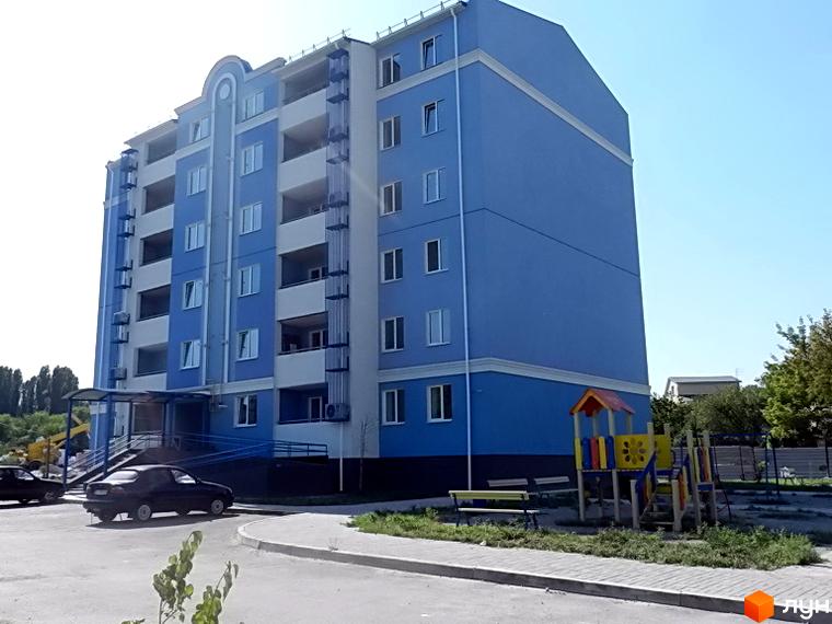 Житловий комплекс Пролісок: багатоповерховий будинок синьо-білого кольору з балконами. Перед будинком розташовані дитячий майданчик та дорога.