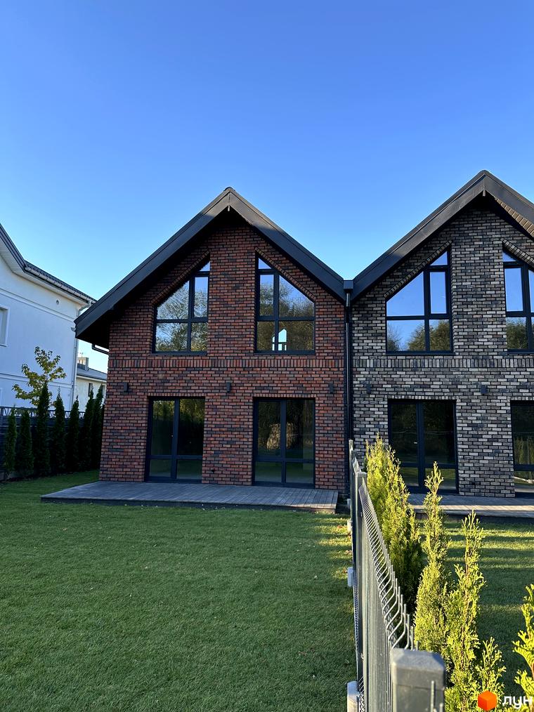 Дуплекси Bavarian Vier з цегляним фасадом та великими вікнами на тлі блакитного неба. Зелений газон та огорожа перед будинками.