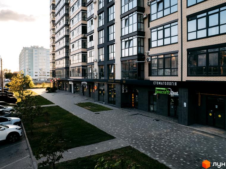 ЖК Династія: будинок з цегли та темних панелей, великі вікна. Комерційні приміщення на першому поверсі, бруковані доріжки, газони.