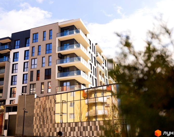 Багатоповерховий будинок Житлового комплексу Grand Hills з різнокольоровими фасадами та скляними балконами. На передньому плані відображення будівлі та дерева.