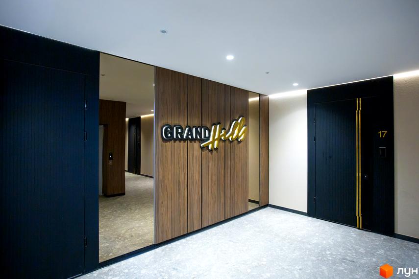 Хол Житлового комплексу Grand Hills з дерев'яними панелями, дзеркалами та підлогою з тераццо. Видно двері до квартир та освітлений логотип Grand Hills.