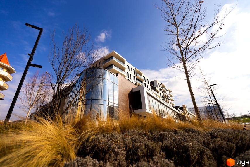 Новобудова Grand Hills з панорамним скляним фасадом та балконами, оточена ландшафтним дизайном з травами та деревами.