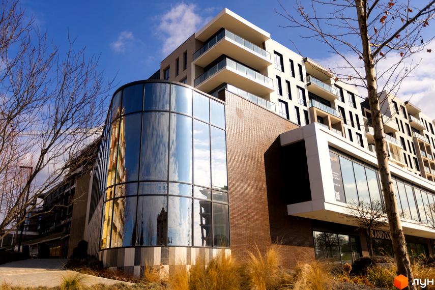 Житловий комплекс Grand Hills з вигнутим скляним фасадом та балконами на верхніх поверхах, оточений деревами та сухою травою.