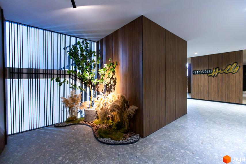 Інтер'єр лобі Новобудови Grand Hills з дерев'яними панелями та логотипом. Простір прикрашає озеленена зона з деревом і травами.