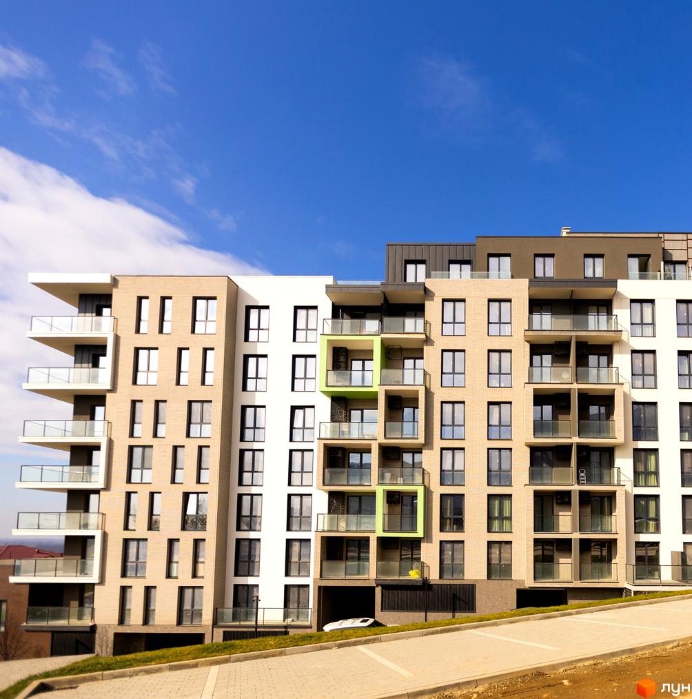 Зовнішній вигляд новобудови ЖК Grand Hills з балконами та вікнами на фоні блакитного неба. Прибудинкова територія з парковкою та зеленню.